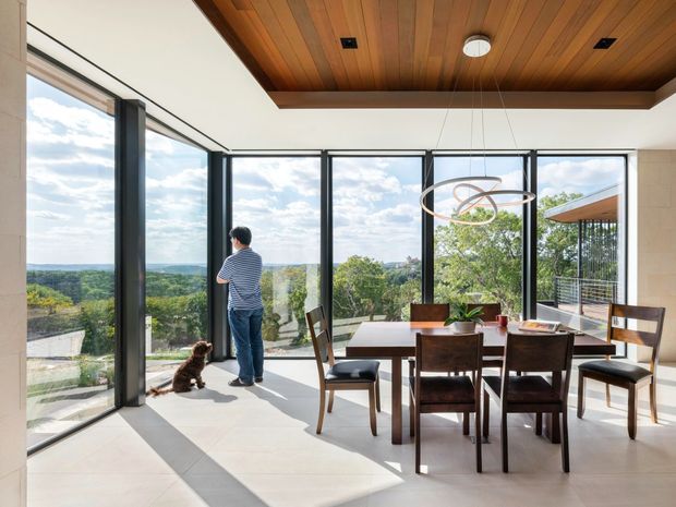 Bright dining area with a timber-lined recessed ceiling, large black-framed floor-to-ceiling glazing, and a dark timber dining table with matching chairs. The space opens to expansive treetop and distant landscape views, with a contemporary pendant light and a small balcony or terrace visible beyond the glazing.