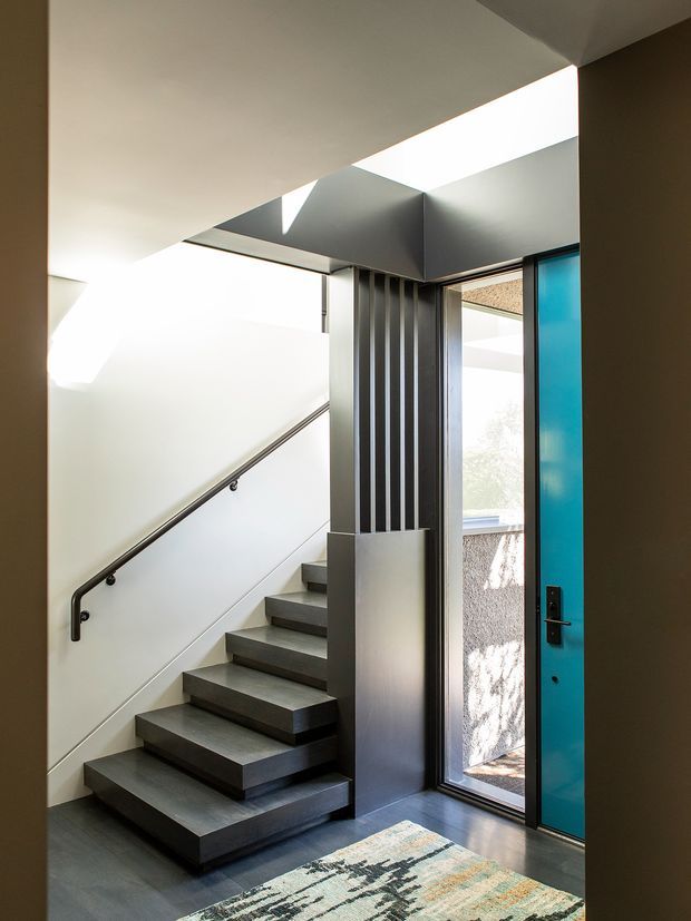 A contemporary residential entryway with a floating staircase leading upward beside a vivid turquoise front door. The stair hall features dark grey stair treads, a slim black handrail, vertical dark metal screening, strong geometric ceiling planes, and a bright skylight that brings in natural light. A patterned rug sits at the entrance, with a glimpse of the exterior visible through the open doorway.