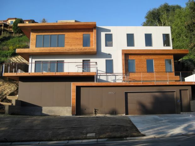 Exterior view of a contemporary multi-level hillside residence with a white rendered upper volume, extensive warm timber cladding, dark metal-framed windows, projecting balconies with slim metal balustrades, and an integrated double garage. The home is set on a steep urban site with neighbouring houses and mature vegetation visible around it.