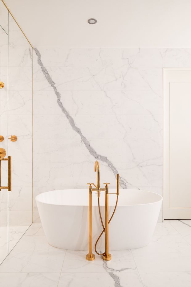 A refined bathroom featuring a freestanding white oval bathtub set against full-height white marble-effect wall and floor tiles with prominent grey veining. Brushed brass/gold bath filler, hand shower, and wall fittings provide a warm metallic accent. A frameless glass shower screen is visible at left, with a white paneled door at right and recessed ceiling lighting.