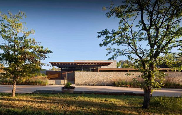 Single-storey contemporary residential building viewed from across a landscaped verge. The low horizontal structure has a broad shallow-pitched roof with exposed timber rafters, dark-framed glazing, and long tan brick or stone masonry walls. Mature trees and planted landscaping frame the facade.