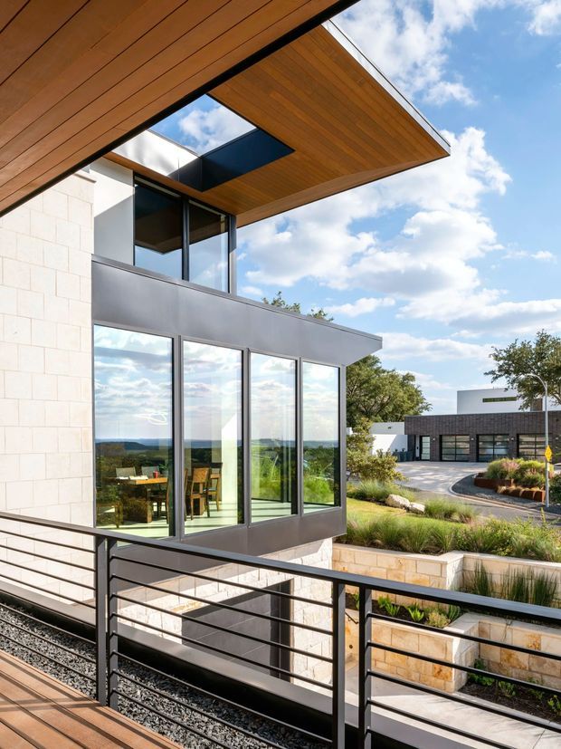 A contemporary multi-level building exterior viewed from a covered terrace or balcony. The facade combines pale rectangular masonry cladding, dark metal-framed floor-to-ceiling glazing, and warm timber-lined projecting soffits with a skylight opening. Black horizontal metal balustrades border the foreground terrace, while landscaped terraces, a paved driveway, and a separate dark-clad building are visible beyond. The image is bright and architectural, with expansive views through the glazing and a partly cloudy sky.