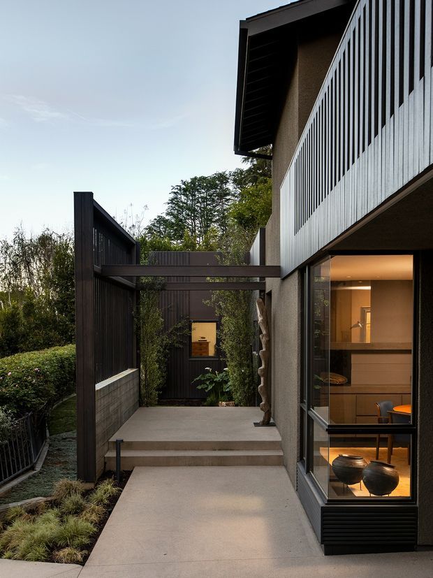 A contemporary residential exterior viewed from a landscaped walkway and terrace. The building features dark vertical cladding and screening, a light rendered wall, broad glazed openings, projecting roof forms, and a prominent upper-level vertical metal balustrade. Concrete steps and paving lead toward a recessed entry courtyard, with warm interior lighting and furniture visible through the glazing.