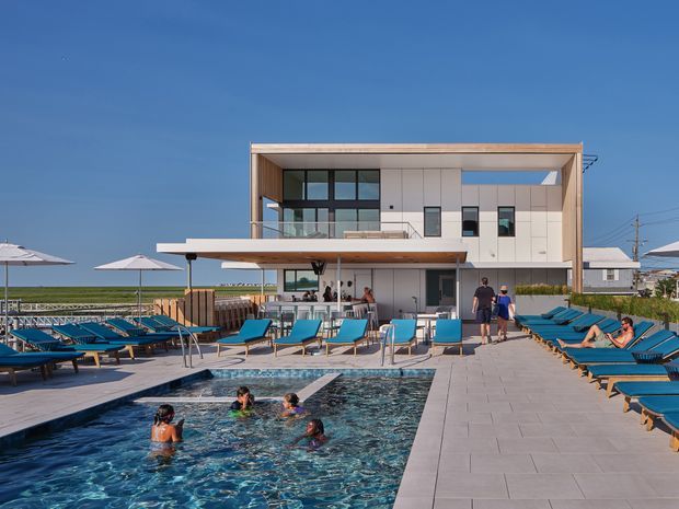 A contemporary multi-level poolside building beside a waterfront, with a rectilinear white facade, extensive glazing, timber-toned soffits, broad flat roofline and projecting terrace. The foreground contains a swimming pool, pale paved deck, blue sun loungers, umbrellas and outdoor seating.