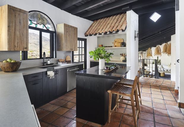 A rustic, Mediterranean-influenced kitchen with dark timber cabinetry, pale stone benchtops, a central island, terracotta tiled flooring, and exposed dark ceiling beams. The kitchen includes an arched glazed window, black-framed doors and windows, a tiled pitched-roof feature over open shelving, and woven bar stools. A connected open area with decorative woven pendant lights is visible beyond the kitchen.