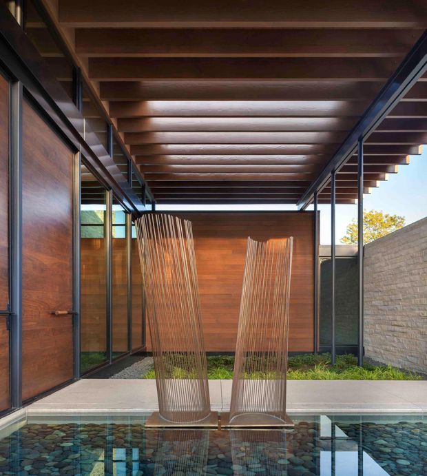 A sheltered architectural courtyard with a shallow reflective water feature in the foreground, two tall woven lounge chairs, dark metal-framed glazing, warm timber wall cladding, exposed timber roof beams, and a textured masonry boundary wall. The composition emphasizes a refined indoor-outdoor connection and strong linear geometry, with a small planted garden visible behind the seating.