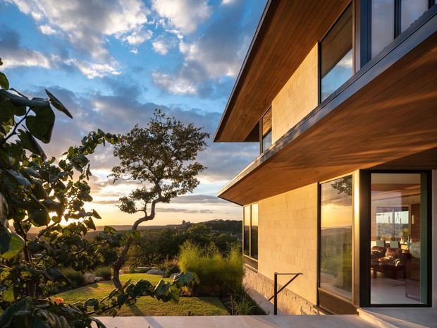 Exterior architectural view of a contemporary two-storey home at sunset, with dramatic cantilevered roof planes, warm timber soffits, large dark-framed windows and glazed doors, and light stone or masonry wall cladding. The house opens toward a landscaped, elevated-looking garden and broad natural landscape beyond.