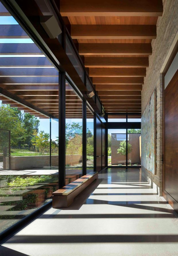 A long, refined interior corridor or gallery with full-height black-framed glazing on one side, exposing a landscaped courtyard and garden. Exposed timber beams and timber ceiling boards run overhead, while a textured stone wall with an inset artwork lines the opposite side. Strong sunlight creates dramatic linear shadows across the pale floor, and a low timber built-in bench runs along the glazing.