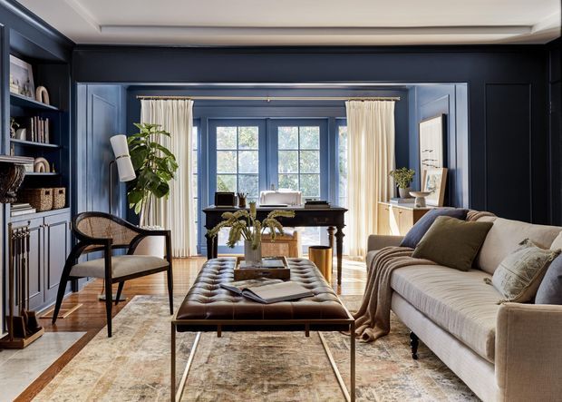 A refined living room with deep navy wall panelling and built-in shelving, a cream upholstered sofa, a tufted leather ottoman, timber flooring, and a large neutral patterned rug. Tall windows with sheer curtains bring natural light into the room, while potted greenery, framed artwork, and carefully styled shelving add warmth. The space has a classic, tailored character with contemporary furnishings.