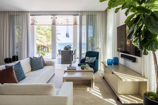 Bright contemporary living room with a large cream sectional sofa, paired upholstered armchairs, layered coffee tables, and a sleek white built-in media console. Floor-to-ceiling sheer curtains frame glazed doors opening to a sunlit balcony and ocean view, while timber ceiling detailing, pendant lights, blue accessories, and abundant indoor greenery add warmth and a coastal atmosphere.