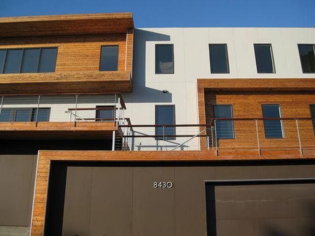 Contemporary multi-storey residential facade with a crisp white rendered central volume, extensive warm timber cladding and projecting balconies. Dark-framed windows, slim metal balcony balustrades, and integrated garage doors create a clean modern street elevation.