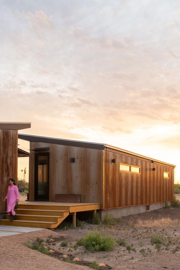 Exterior photograph of a compact single-storey modern dwelling with a long, low-pitched roof and extensive vertical timber cladding. The home is raised slightly above a sparse, dry landscape, with a small timber entry deck and broad steps leading to a dark glazed entrance. Narrow horizontal windows and warm sunset light emphasize the simple linear form.