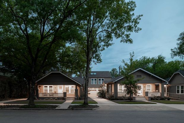 Street-level exterior view of a small residential development consisting of several closely spaced single-storey gable-roof homes, with front porches, light-coloured siding, dark trim, paved driveways and mature street trees. A taller two-storey dwelling is visible behind the front homes.
