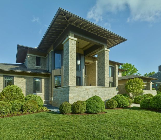 Exterior view of a substantial two-storey residence with pale stone masonry walls, dark-framed glazing, broad projecting roof eaves, and a prominent covered entry porch supported by square stone columns. The landscaped front yard features manicured lawn and rounded clipped shrubs under a bright blue sky.