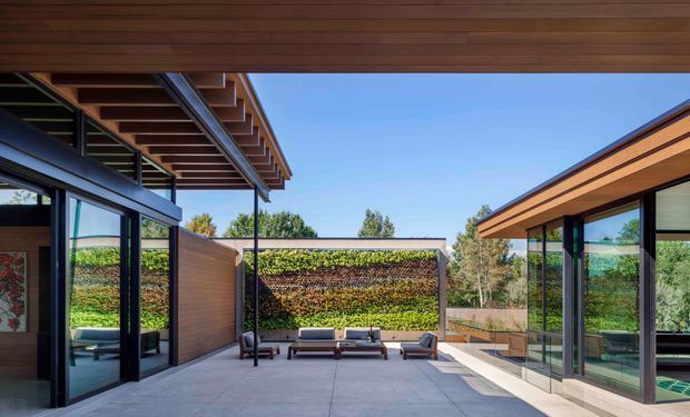 A contemporary courtyard between two low-profile glazed wings, with broad overhanging timber-clad roofs, exposed structural rafters, dark-framed floor-to-ceiling glass, and a paved outdoor seating area. A planted vertical garden forms the rear boundary, while the open sky and landscaping create a bright, private indoor-outdoor setting.