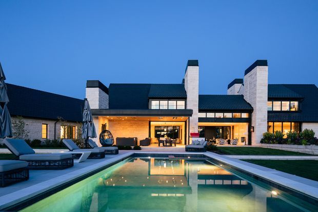 A large upscale multi-wing residence photographed at dusk from the landscaped pool area. The home has dark pitched roofs, pale masonry chimney towers, extensive warm-lit glazing, and a broad covered outdoor entertaining area opening toward a rectangular swimming pool. Poolside loungers, umbrellas, built-in planting and manicured lawn surround the water.