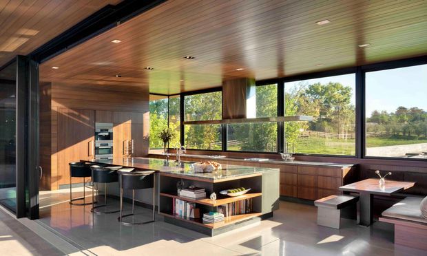 A spacious contemporary kitchen and dining area with extensive floor-to-ceiling glazing, dark framed windows, warm timber-lined ceiling and walls, a large stone island with open shelving, integrated appliances, bar stools, and an adjacent built-in dining nook. The room opens toward a green outdoor setting and is filled with natural light.