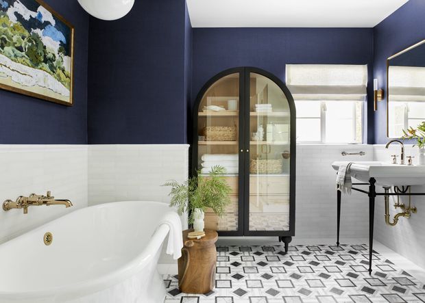 A bright, refined bathroom with deep navy walls, white half-height wall tiling, a freestanding white bathtub, patterned geometric floor tiles, and a white pedestal-style basin with exposed brass plumbing. A black-framed arched glass cabinet provides storage, while a large mirror and window enhance the light. Brass fixtures and a potted plant add warmth and character.