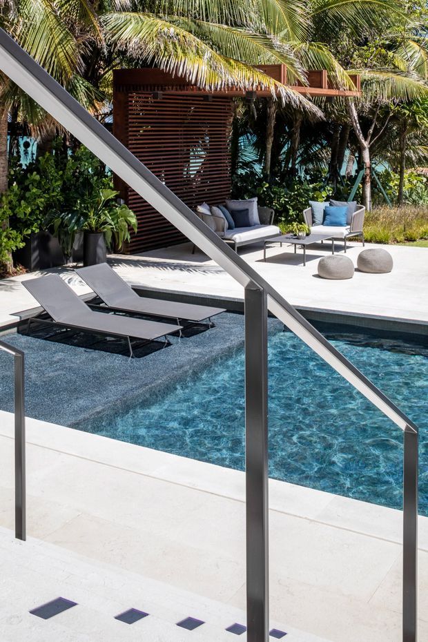A bright tropical pool courtyard with a rectangular swimming pool, pale paved surrounds, dark in-water loungers, and a modern metal handrail in the foreground. A shaded outdoor lounge beneath a timber slatted structure sits among dense palms and tropical planting, with neutral seating, blue cushions, and rounded ottomans.