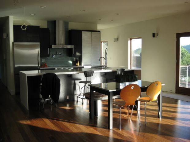 Open-plan kitchen and dining area with dark cabinetry, stainless-steel appliances, a long island with bar stools, and a glass-topped dining table with orange chairs. Warm timber flooring, recessed ceiling lights, and large windows and glazed doors bring in strong afternoon sunlight and views of the surrounding landscape.