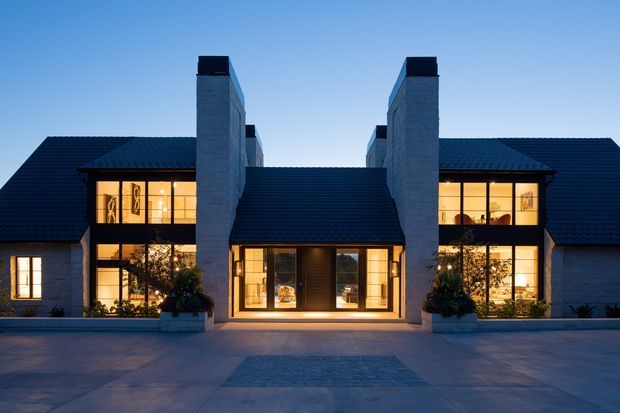 Symmetrical contemporary residential facade photographed at dusk, with dark pitched roofs, pale masonry walls, prominent paired chimneys, and warm light glowing through large windows and the central glazed entry. A broad paved forecourt and formal planters frame the building.