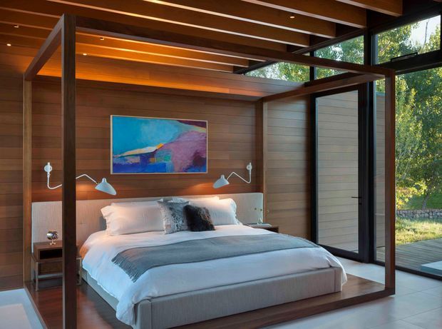 A warm, modern bedroom with a low platform bed framed by dark timber posts and exposed timber ceiling beams. Horizontal timber wall panelling continues behind the bed, with integrated bedside shelves and wall-mounted reading lights. Large black-framed glazed doors and clerestory windows open to a leafy outdoor setting, while a colourful abstract artwork provides an accent above the upholstered headboard.