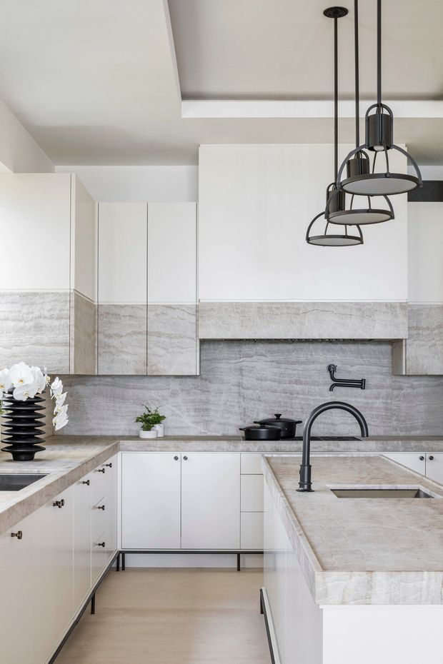 A refined contemporary kitchen with white flat-panel cabinetry, pale stone slab benchtops and matching full-height splashback, and matte black plumbing fixtures and hardware. A large island with an undermount sink occupies the foreground, while three clustered black pendant lights hang above. The kitchen has a clean U-shaped layout, integrated storage, a concealed rangehood or cooking zone, and warm pale flooring.