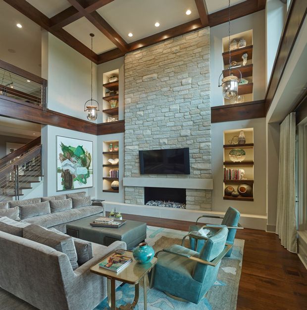 A spacious double-height living room with a large stone-clad fireplace wall, built-in shelving, a wall-mounted television, exposed dark timber ceiling beams, and an open mezzanine level. A large sectional sofa, teal armchairs, ottomans, and a coffee table sit on a patterned rug, with extensive glazing along one side.