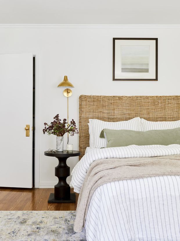 A softly styled bedroom with a woven rattan headboard, white-and-grey pinstriped bedding, a sage green accent pillow, and a textured neutral throw. The room has white walls with decorative trim, warm timber flooring, a black pedestal side table, a brass wall lamp, framed artwork, and a partially open white door.