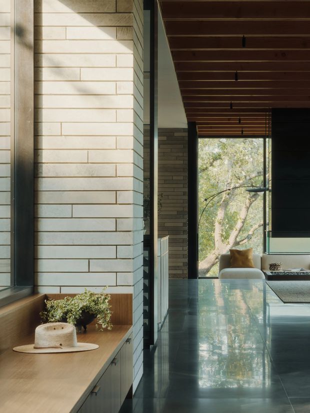 A refined modern interior view focused on a long built-in timber bench and cabinetry beside a large window, with pale horizontal masonry wall tiles, dark metal-framed glazing, a polished dark floor, and a timber slatted ceiling. The space opens toward a seating area and leafy outdoor view, with strong natural light and restrained furnishings.