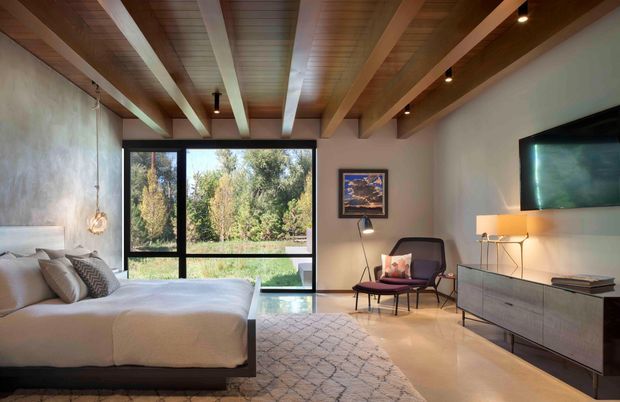 A warm, contemporary bedroom with a low-profile upholstered bed, textured bedding, mid-century-inspired lounge chair, wall-mounted television, and streamlined media console. Exposed timber ceiling beams and timber ceiling boards contrast with pale plaster walls and a polished light-toned floor. Full-height black-framed glazing opens the room visually to a leafy landscape, while layered lighting and neutral textiles create a refined, inviting atmosphere.