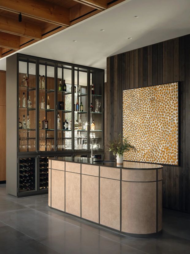 A sophisticated home bar with a curved, paneled bar island, dark timber wall cladding, glass-fronted liquor and glassware storage, integrated wine racks, and a patterned illuminated artwork panel. The space has polished concrete or large-format grey flooring, a white recessed ceiling, black metal framing, and warm timber ceiling detailing.