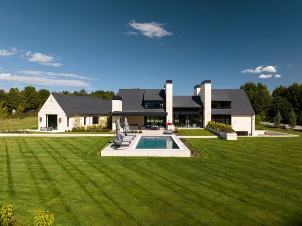 A large, symmetrical residential house viewed from the landscaped rear grounds. The low, sprawling stone-clad home has dark pitched roofs, multiple gabled roof sections, prominent pale masonry chimneys, extensive glazing, and a formal lawn leading to a rectangular swimming pool and paved terrace.