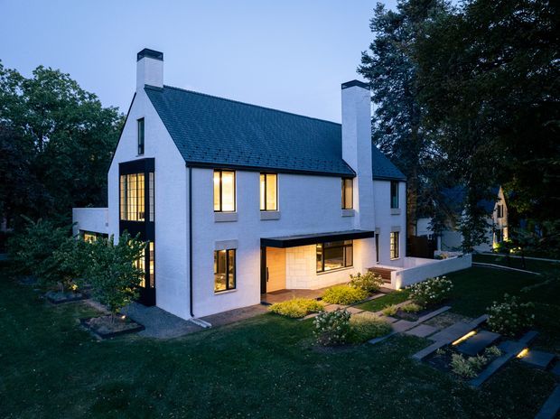 Twilight exterior view of a substantial two-storey white-rendered house set within a landscaped garden. The home has a steep dark pitched roof with prominent white chimneys, tall black-framed windows, warm interior lighting, and a recessed ground-floor entry or living area beneath a projecting upper floor. Paved pathways, planting beds, lawn, and mature trees surround the building.