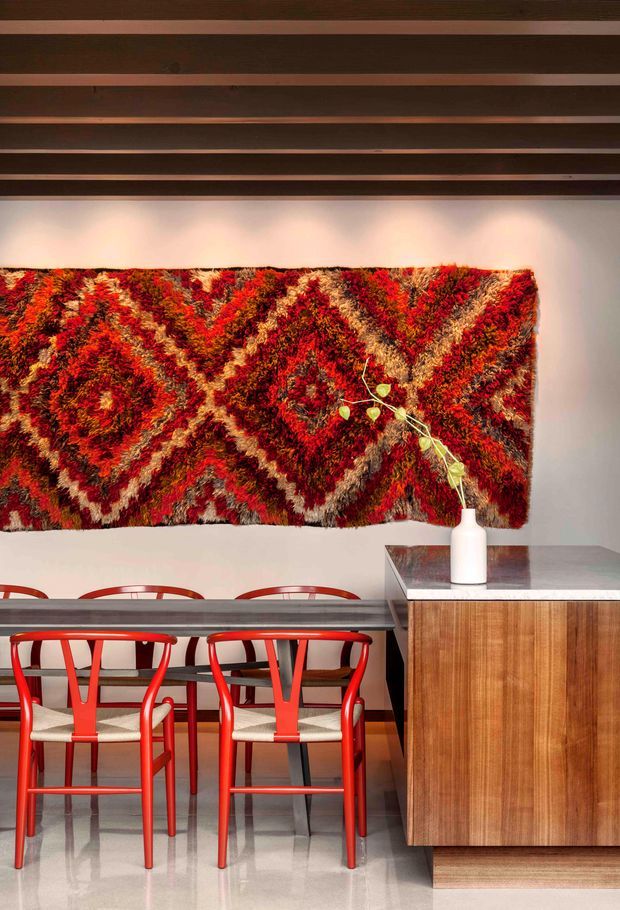 A richly styled dining area with a long dark dining table and vivid red wooden chairs with woven seats. A large red, orange, cream, and brown geometric textile wall hanging forms the focal point above the table. The room has exposed dark timber ceiling beams, pale walls, a polished light floor, and a fixed timber sideboard with a grey stone top and a white ceramic vase.