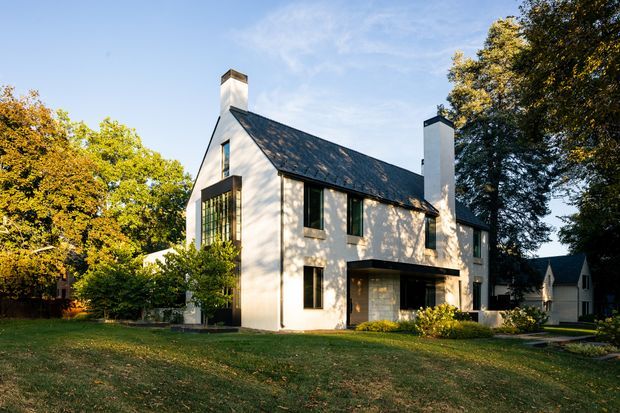 Exterior view of a refined two-storey residential house set within a mature, leafy suburban garden. The home has pale rendered walls, a steep dark pitched roof, prominent chimneys, tall black-framed windows, and a glazed ground-floor corner extension or sunroom. Warm late-day sunlight highlights the facade and surrounding trees.