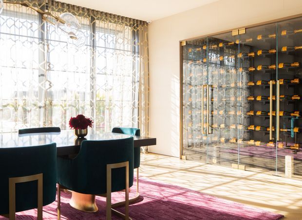 A bright, refined dining area with a dark rectangular dining table and deep teal upholstered dining chairs, positioned beside expansive patterned glazing. A dramatic glass-fronted wine storage wall with extensive bottle racks and brass-toned hardware dominates the right side. Sunlight casts intricate shadows across the pale floor and walls, while a large magenta-purple rug and floral centerpiece add bold colour. The space has an elegant, glamorous character with strong decorative detailing in the window treatment and cabinetry.