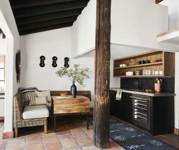 A rustic-modern open-plan kitchen and sitting area with white walls, exposed dark timber ceiling beams, a prominent rough-hewn timber support post, terracotta tiled flooring, and black cabinetry. The kitchen features open timber shelving, a glossy dark tiled splashback, a dark benchtop, and integrated wine storage. A woven lounge chair, timber console, cushions, and simple black wall sconces create a warm, textural interior.