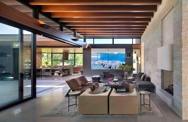 Spacious contemporary open-plan living room with a double-height-feeling timber-beamed ceiling, extensive black-framed glazing, and a large natural stone feature wall. The room includes a conversation seating arrangement, fireplace or media feature, polished concrete flooring, and views toward a landscaped exterior. Warm timber, stone, glass, and neutral upholstery create a refined indoor-outdoor atmosphere.