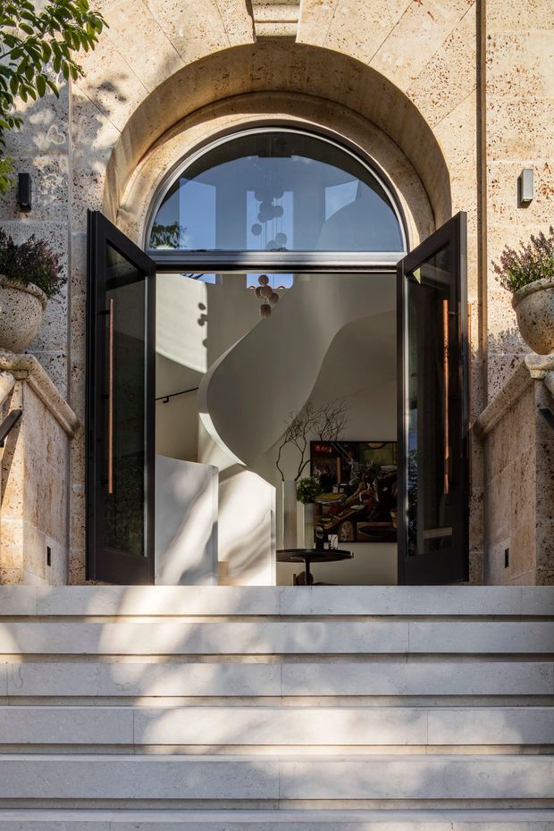 A grand arched entrance viewed from outside, with open dark-framed glazed doors set within a richly textured warm stone facade. Broad pale stone steps lead up to the doorway, while the interior beyond reveals a sculptural curved white stair or wall, a small table, artwork, and decorative lighting. Potted greenery and strong dappled sunlight add an elegant, refined atmosphere.