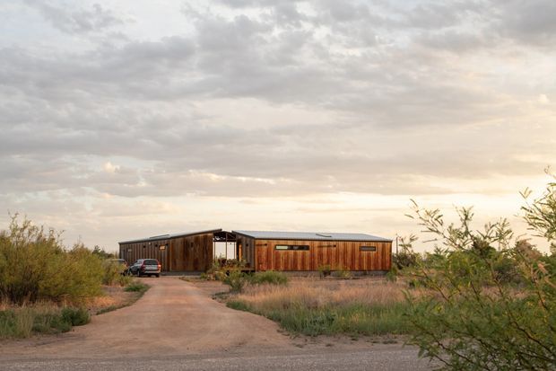 Low, single-storey modern building set within a dry, scrubby landscape. The long rectangular structure has warm vertical timber cladding, dark metal framing and a shallow low-pitched roof, with a central covered entry or breezeway and narrow horizontal windows. A dirt driveway approaches the building, with sparse native-looking vegetation surrounding it beneath an overcast evening sky.