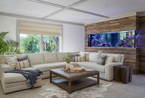 A bright, spacious living room with a large cream sectional sofa, textured neutral cushions, a substantial wooden coffee table, and a pale area rug. A dramatic built-in aquarium is set into a horizontally clad timber feature wall, while large windows with roller blinds overlook lush greenery. The ceiling features recessed lighting and subtle linear detailing.