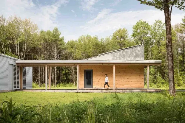 A single-storey contemporary pavilion-style house set within a wooded landscape. The low, elongated structure has a shallow asymmetrical pitched roof, extensive open covered outdoor circulation, warm horizontal timber-clad walls, dark-framed glazing and doors, and a pale grey metal-clad side volume. A broad lawn and mature trees surround the building.