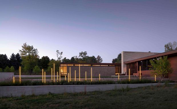 A contemporary low-rise building exterior photographed at dusk. The composition shows a long, dark glazed and solid facade with warm linear lighting, a projecting roof or canopy, and a taller masonry volume to the right. Landscaped planting, concrete retaining walls, and slender illuminated posts define the foreground and approach.