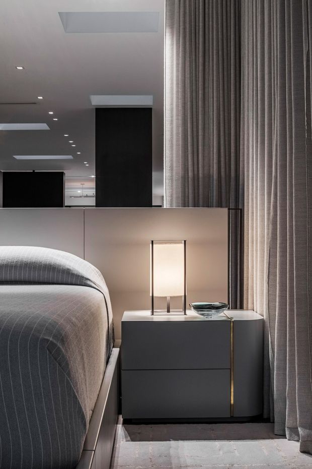 A sophisticated modern bedroom vignette featuring a partially visible upholstered bed, dark minimalist bedside cabinetry, a sculptural table lamp, and a small glass bowl. Full-height textured grey curtains cover the window, while a reflective partition or mirror behind the bed adds depth and reflects recessed ceiling lighting. The palette is restrained in charcoal, grey, taupe, and soft white, creating a refined, calm atmosphere.