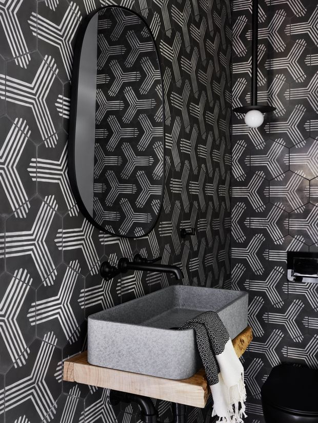A dramatic compact bathroom featuring a grey vessel basin on a light timber wall-mounted vanity, matte black wall-mounted tapware, a large rounded-rectangle black-framed mirror, and a black-and-white geometric patterned wall finish. A sculptural black pendant with a white globe adds a decorative lighting accent. The space has a bold monochrome, graphic aesthetic with warm timber contrast.