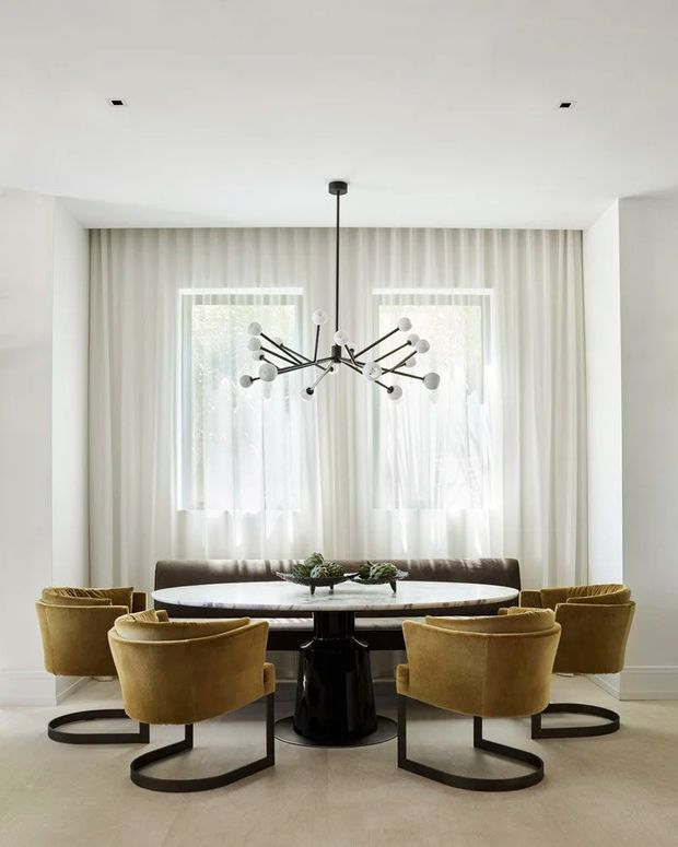 A refined dining room with a large oval dining table surrounded by mustard-gold upholstered barrel chairs. The room features white walls and ceiling, tall windows with sheer curtains, pale flooring, dark table and chair bases, and a sculptural black-and-white multi-arm pendant light. The overall composition is bright, symmetrical, and sophisticated.