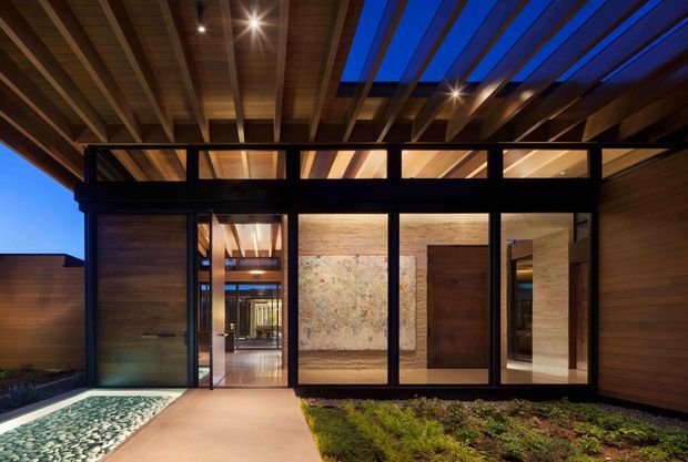 Twilight exterior view of a contemporary architectural entryway. A broad timber-lined soffit with exposed beams and blue accent lighting projects over a glazed entrance facade framed in dark metal. Warm interior light reveals timber ceilings, stone or textured feature walls, and a substantial timber pivot door. A concrete path leads through a landscaped garden toward the entry.