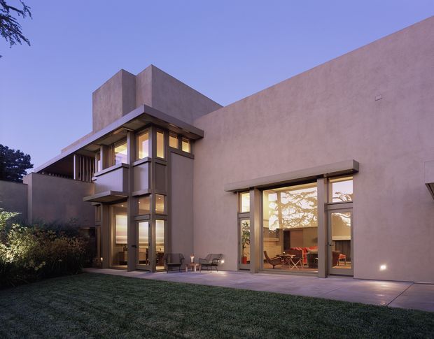 Exterior dusk view of a modern, low-profile residence with a sculptural flat-roof form, smooth warm-toned rendered walls, extensive floor-to-ceiling glazing, and dark metal-framed windows and doors. A glazed projecting entry volume and sheltered terrace connect to a lawn and landscaped garden, with warm interior lighting visible through the windows.
