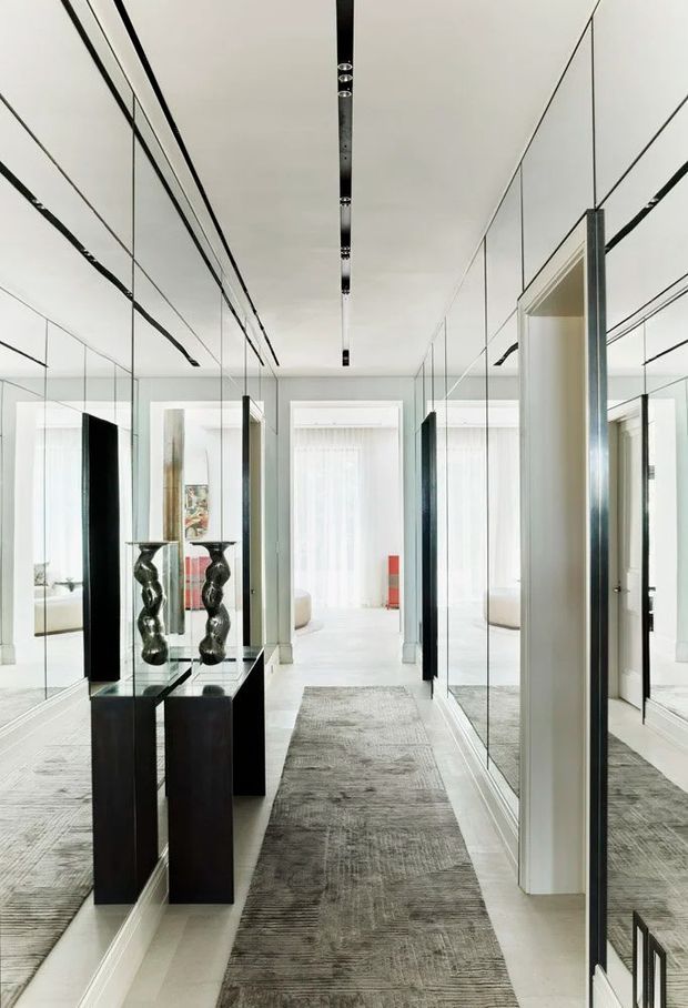 Bright, highly refined hallway or entry corridor lined with full-height mirrors and slim black-framed mirrored doors. A long grey textured runner leads toward a sunlit living area, while two black sculptural pedestal tables with metallic decorative vessels form a central display. The space has crisp white walls and ceiling, concealed linear lighting, and strong symmetrical perspectives created by the mirrored surfaces.